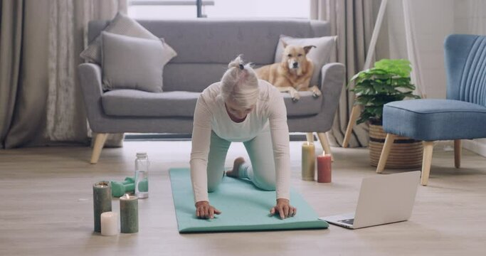 Mature woman practicing yoga while watching an online tutorial video on her laptop at home. Female yogi stretching on an exercise mat in a virtual fitness class, finding balance and inner peace
