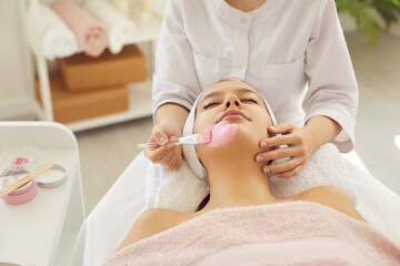 Obraz na płótnie Canvas Woman getting facial treatment. Happy relaxed beautiful young lady lying under soft towel on spa bed in beauty salon or spa center and enjoying pink kaolin clay mask for fresh, clear skin, close up