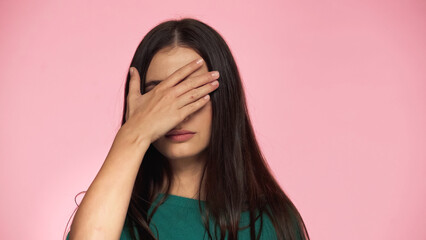 brunette young woman in green blouse covering eyes with hand isolated on pink.
