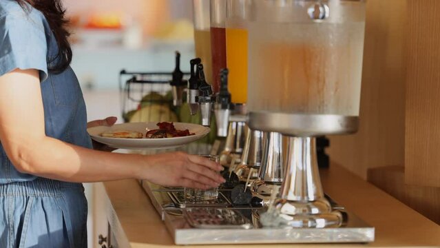 Close up of woman hand pulling down the lever of juice dispenser for fresh orange juice and holding plate of Fried eggs, Bacon, standing near row of fresh juice at buffet restaurant or hotel.
