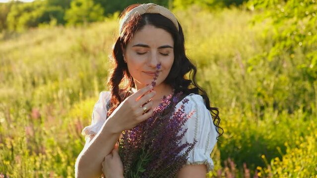 A young woman in a dress stands in a meadow with purple flowers in her hands at sunset. Evening walk in the summer on the meadow. Summer travel.