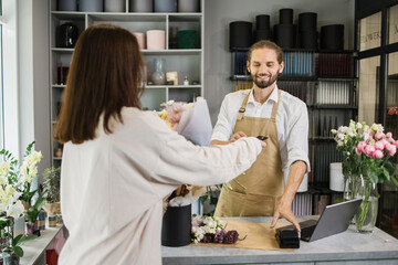 Young bearded florist male receiving discount card from regular female customer and making payment at terminal in flower shop.