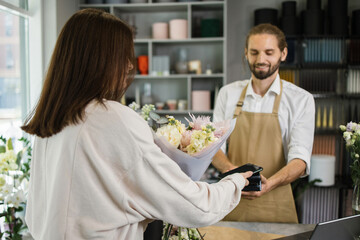 Fototapeta na wymiar Caucasian female using electronic credit card on her smartphone paying for purchase using payment terminal in flower shop. Client buying bouquet making payment in store