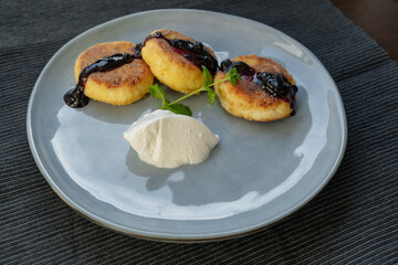 Delicious fried cheesecakes with a crispy crust, poured with cherry jam, served on a gray ceramic plate with a ball of sour cream and decorated with a sprig of fragrant mint
