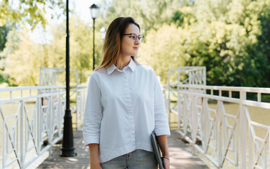 Pretty young woman wearing white shirt, jeans and eyeglasses holding laptop standing on bridge in park and looking away. Portrait of waist-length caucasian female student or freelancer outdoors