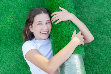 Smiling young caucasian girl lying on the lawn and hugging a new roll of artificial turf. Soft and squishy artificial grass ready to lay