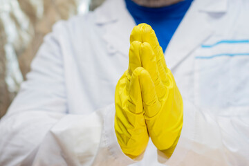 A doctor in a medical gown and protective gloves holds folded palms in front of him. The face is not visible. Gesture of greeting, gratitude or prayer.