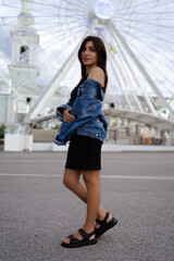 Naklejka premium Stylish beautiful young brunette woman model in summer , dressed in a black dress and jeans jacket. Full body model, vertical photo , ferris wheel in the background. Street fashion. 