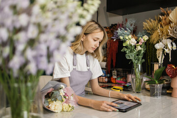 Portrait of attractive woman florist working on tablet at workplace. Concentrated female sitting at...