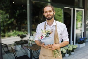 Attractive man inviting to his flower shop. Attractive smiling young florist in apron . Smiling man holding bouquet outside of flower shop.