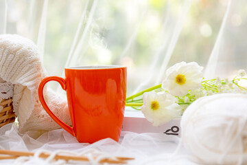 Coffee morning.  Orange steaming cup of hot coffee for relax after working hobbies crochet on white wooden near window with flowers