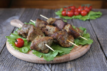 The pieces of grilled pork on a wooden  skewers are on the cutting board with tomatoes and greens. 