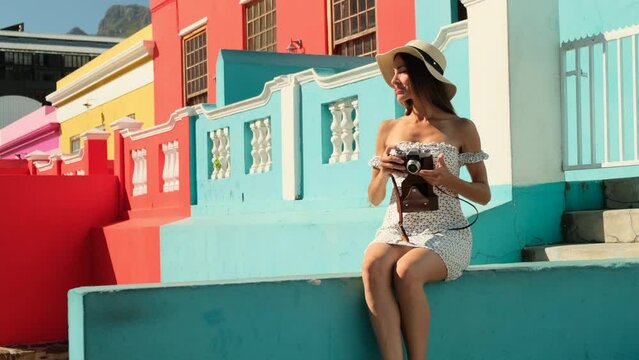 an elegant girl with beautiful legs in a dress sits on a wall near colorful houses in italy and makes a photo. girl tourist in a dress and hat takes pictures of the city with a film phone on vacation