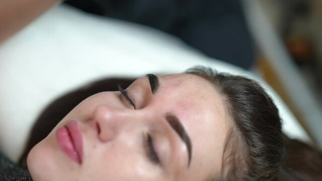 Face of young gorgeous woman with beautician hands tattooing second eyebrow with microblading pen. Beautiful Caucasian slim client with closed eyes in beauty salon doing permanent makeup