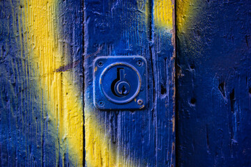 Old-styled front door and metal lock, padlock view. A blue and yellow weathered rusty wooden painted gates. Wooden slats, boards, surface. Entrance door to an old house, flat. Architectural details.