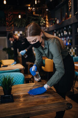Young restaurant waiters cleaning and disinfecting tables and surfaces during Coronavirus pandemic disease. They are wearing protective face masks and gloves.