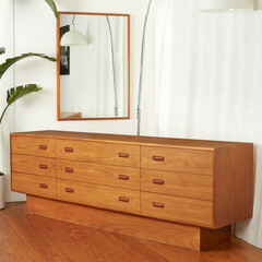Beautifully crafted Scandinavian Modern design dresser. Wooden Mid-century modern style furniture. Angled view with mirror and houseplant to the side and white curtains in the background. 