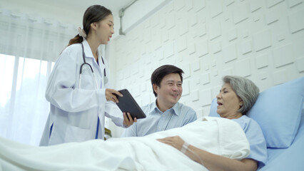 Obraz premium Asian man sitting beside his elder mother on the bed in hospital ward and doctor is coming to tell him about the medical analyze result.