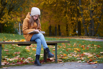 Young stylish teenage rgirl sitting on the bench in the autumn park and reading book.