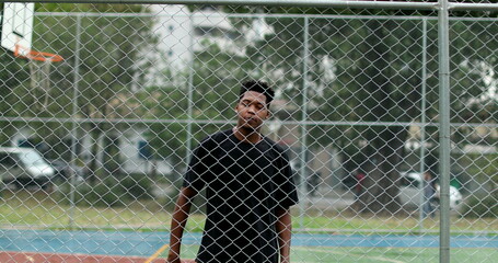 Young black man behind metal fence at basketball court