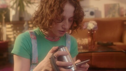 Tilting up of ginger Caucasian girl in outfit in 80s fashion sitting on floor in living room at daytime, using old-fashioned video camera