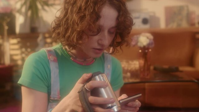 Tilting up of ginger Caucasian girl in outfit in 80s fashion sitting on floor in living room at daytime, using old-fashioned video camera
