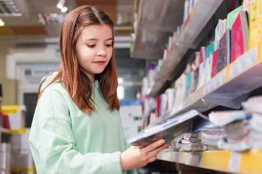 schoolgirl looking at new copybook near blurred rack in stationery shop.