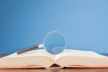 open book with magnifying glass on wooden desk in information library of school or university, concept for education,reading , study, copy space and blue background.