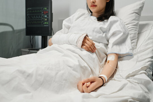 Horizontal shot of young Asian woman with arm in cast lying on hospital bed in emergency room getting intravenous therapy