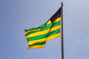 Uma bandeira do estado de Goiás hasteada e balançando ao vento com céu ao fundo.