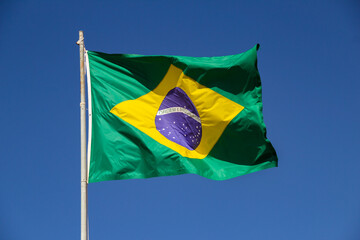 Uma bandeira do Brasil balançando ao vento  com céu azul ao fundo.