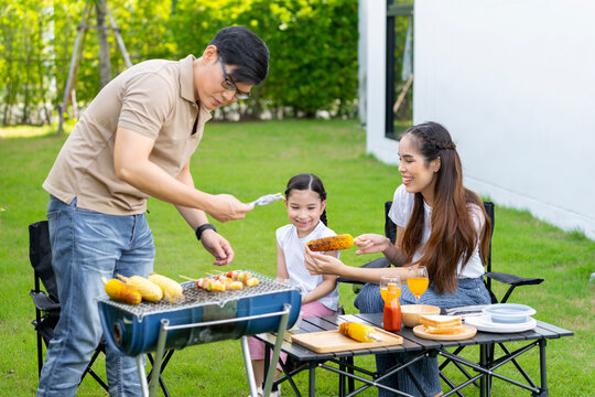 An Asian Family Of Parents And Daughters Camping With BBQ Tires. In The Front Yard Outside The House Together Happily