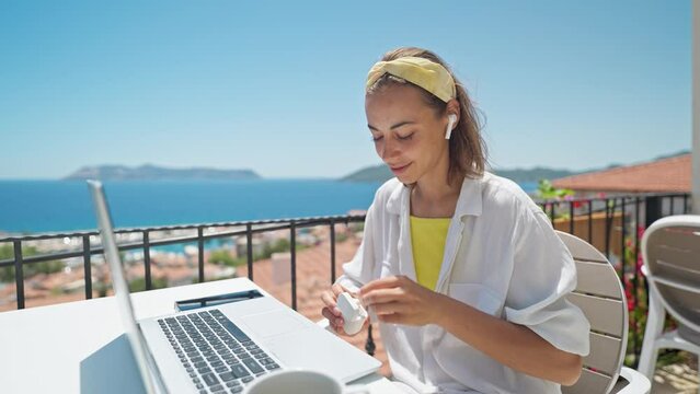Joyful attractive woman wearing earphones, looking at laptop computer while sitting at outdoor cafe during Mediterranean vacation