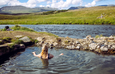 Woman relaxing and bathing in natural geothermal heated hot pool, hot spring in Iceland in summer. Green rolling hills and blue sky on background. Hot spring is called Fosslaug hot pot.