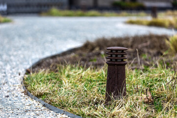 A garden ground lamp made of iron mounted in grass near gravel pathway. Small architectural forms.