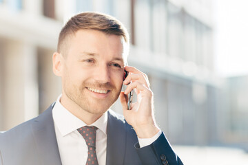 Confident happy middle aged businessman communicates via cell phone, discusses financial report with business partner, wears formal clothes, stands outdoor against blurred background. Busy CEO