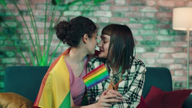 In living room on the sofa closeup to the camera very lovely lesbian couple hugging closeup each other and holding in hands LGBT rainbow flag. Shot on ARRI Alexa Mini