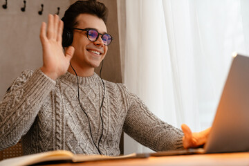 Young white man wearing eyeglasses smiling while using laptop and headphones in cafe