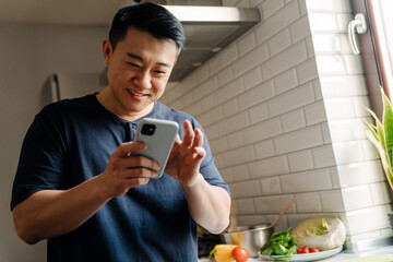 Adult brunette asian man smiling and using cellphone at home
