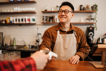 Adult asian man holding payment terminal while working in cafe indoors