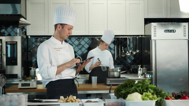 Restaurant kitchen: portrait of male and female cooks. Male Chef checks the availability of products in the warehouse using a tablet