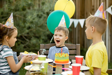 Cute funny four year old boy celebrating his birthday with family or friends with homemade baked cake in a backyard. Birthday party for kids. His gets presents gift box.