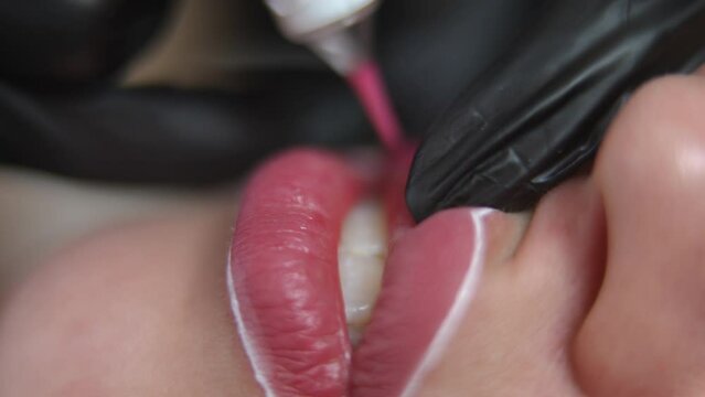 Close-up female lips with white contours and blurred beautician hand tattooing at background with microblading pen. Unrecognizable Caucasian young confident woman doing permanent makeup