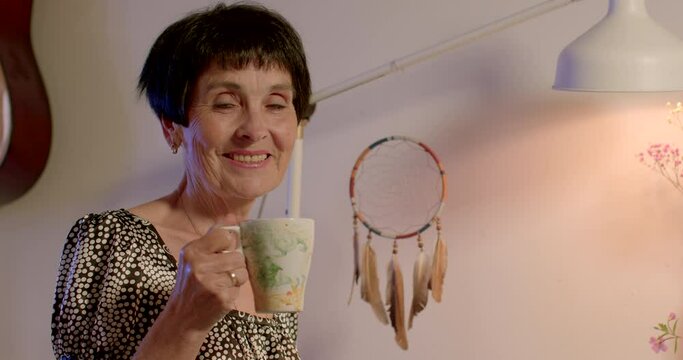 Portrait of a senior woman with a mug of hot drink in her hands. She is looking at the camera, smiling. Goodbye waiting. A dream catcher hangs in the background.