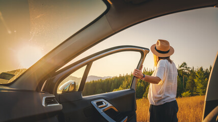 Young woman traveler in hat getting out of car on sunset field during auto travel