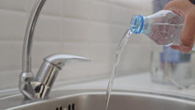 A close-up of a man's hand pours water from a plastic bottle into a home metal sink against a white wall.