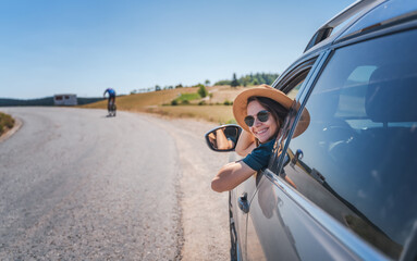 Happy young woman driver traveler in hat and sunglasses sitting behind the wheel of car on summer country road
