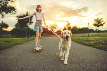 Portrait of teenage girl petting golden retriever outside in sunset running on her longboard with dog