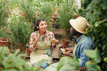 Young Black man and Hispanic woman sitting relaxed in greenhouse having lunch and talking about...