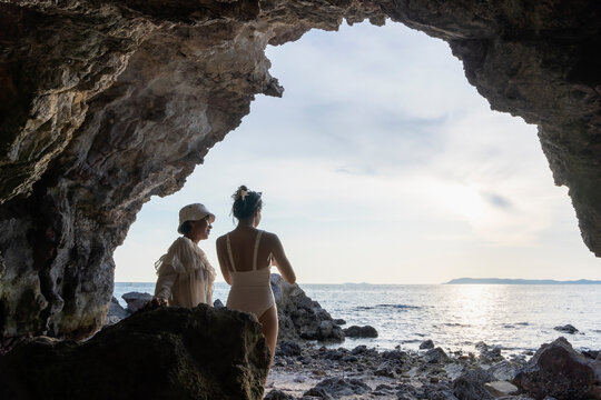 Two women taking photos in cave overlooking sea at sunset or sunrise. Mother and daughter relaxing on tropical beach travel summer holidays. Familly tourists enjoy traveling to exotic nature.